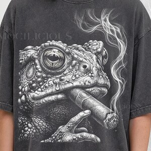 May include: Dark gray t-shirt featuring a detailed illustration of a toad smoking a cigar. The toad's eye is in focus, with smoke rising from the cigar. The text "MOCHILICIOUS" is at the top, and "PEPPER COMFORT COLORS" is at the bottom.