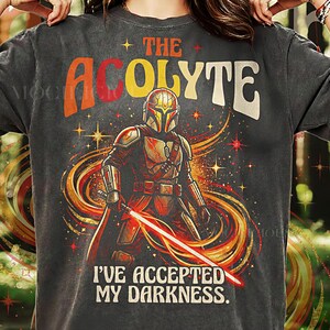 Könnte beinhalten: Dunkelgraues T-Shirt mit einer Grafik eines Mandalorianer-Charakters mit rotem Lichtschwert und dem Text "THE ACOLYTE" und "I'VE ACCEPTED MY DARKNESS." Das Design enthält orange, gelbe und rote Akzente mit Sternendetails.