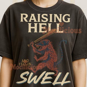 Könnte beinhalten: Dunkelgraues T-Shirt mit dem Text "RAISING HELL" über einer Cartoon-Ratte mit einem Baseballschläger. Unter der Ratte steht der Text "AND FEELIN' SWELL". Die Ratte ist rot und blau.