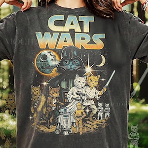 Może przedstawiać: Ciemnoszara koszulka z grafiką przedstawiającą koty w parodii Gwiezdnych Wojen. Projekt zawiera napis "CAT WARS" w kolorze niebieskim i pomarańczowym, z różnymi postaciami kotów w kultowych rolach, na tle gwiezdnego nieba.