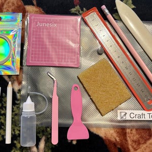 Puede incluir: Un conjunto de herramientas y suministros para manualidades, que incluye una esterilla de corte rosa, una regla, un lápiz y varias herramientas pequeñas. La imagen también muestra pedrería, un frasco de pegamento y pequeños lazos decorativos. El texto "Craft Tools" es visible.