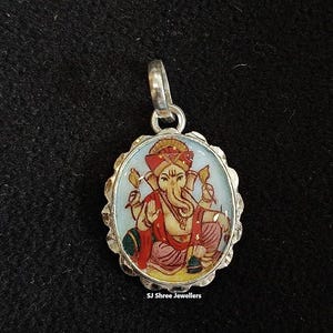 May include: Oval silver pendant featuring a colorful illustration of the Hindu deity Ganesha. The pendant has a scalloped edge and a bail for a chain. The image of Ganesha is in red, yellow, and blue. The text "SJ Shree Jewellers" is at the bottom.