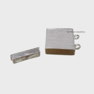 May include: Two silver-colored jewelry pieces. One is rectangular, and the other is a square with a textured surface and brown base. The square piece has two small loops. The text "SJ Shree Jewellers" is visible on the square piece.