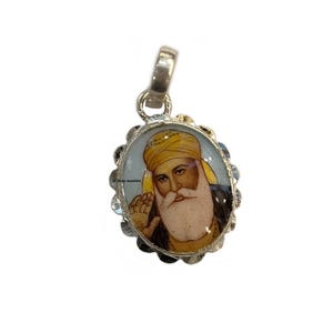 May include: Oval silver pendant featuring a religious figure with a yellow turban and white beard. The pendant has a detailed image and a decorative, scalloped border. The bail is silver.