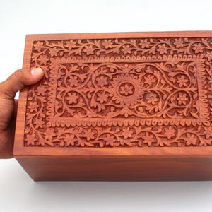Puede incluir: Una caja de madera rectangular con intrincadas tallas florales en la tapa. La caja es de color marrón cálido y parece estar sujeta por una mano. Las tallas presentan flores detalladas y patrones arremolinados.