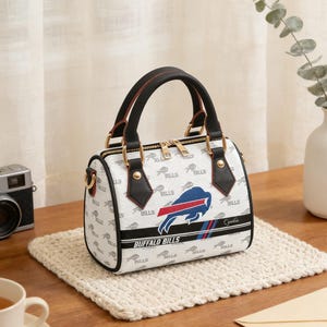Può includere: Borsa a mano bianca e nera dei Buffalo Bills con manico superiore e tracolla staccabile. La borsa presenta il logo della squadra e la scritta "Buffalo Bills". La borsa è appoggiata su un sottobicchiere intrecciato.