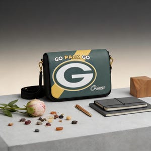 Peut inclure: Sac à bandoulière vert et noir avec le logo des Green Bay Packers et l'inscription "GO PACK GO". Le sac a une bandoulière noire et des accessoires dorés. Le sac est posé sur une surface grise avec un stylo, des carnets et une fleur.