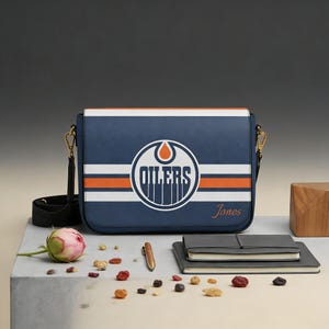 Op de afbeelding: Een blauwe messenger bag met het Edmonton Oilers-logo, witte en oranje strepen en de naam "Jones". De tas heeft een zwarte riem en goudkleurige hardware. Een roze bloem, pen en notitieboekjes zijn ook zichtbaar.