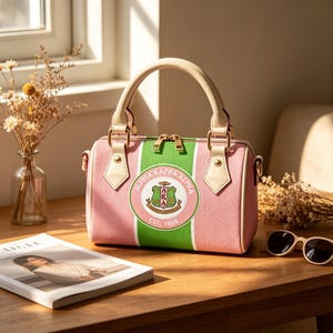 May include: Pink and green Alpha Kappa Alpha handbag with a cream-colored handle and accents. The bag features a circular emblem with the sorority's name and crest. The bag has a gold zipper and a detachable shoulder strap.