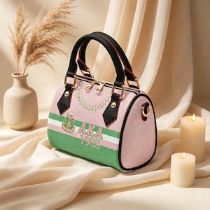 May include: Pink and green barrel-shaped handbag with black handles and gold hardware. The bag features a pearl necklace design and the text "AKA 1908" with a crest. The bag is set against a soft, neutral background with candles and a vase.