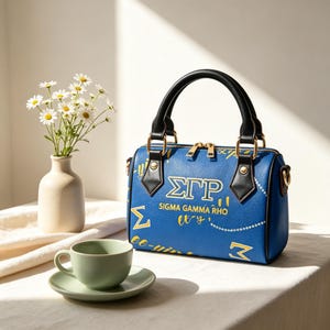May include: Blue handbag with black handles and gold hardware. The bag features the Greek letters ΣΓΡ in gold lettering, along with the words "Sigma Gamma Rho". A small vase with daisies and a teacup are also in the image.