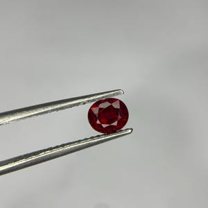 May include: A vibrant, oval-cut red gemstone held by a pair of silver-toned metal tweezers. The stone features a faceted surface that reflects light, showcasing a deep, saturated crimson hue throughout its transparent, polished crystalline structure.