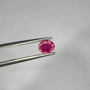 Peut inclure: Une pierre précieuse rubis de taille ovale avec une teinte rouge rosée profonde et de multiples facettes qui reflètent la lumière. La pierre est maintenue fermement entre les deux pointes métalliques dentelées d'une pince argentée.