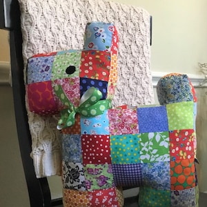 May include: A handmade patchwork dog-shaped pillow. The pillow is constructed from various fabric squares in a vibrant mix of colors and patterns, including florals, polka dots, and geometric designs. A green ribbon is tied around the neck. The pillow is approximately 12 inches tall.