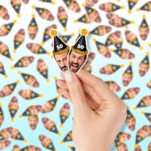 May include: Two stickers featuring a man's face wearing a black party hat with the number 50. The stickers are held in front of a background of champagne bottles. The party hats have a gold pom-pom on top.