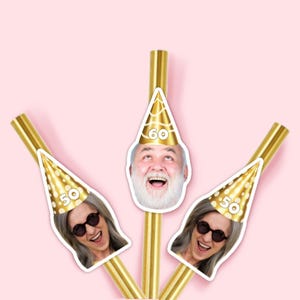 May include: Gold-colored straws with attached cutouts of smiling faces wearing party hats. The hats display the numbers "50" and "60" on a pink background. The straws are arranged in a fan shape.