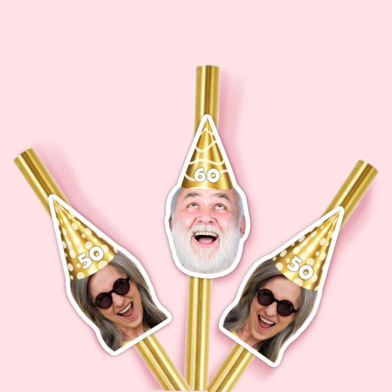 May include: Gold-colored straws with attached cutouts of smiling faces wearing party hats. The hats display the numbers "50" and "60" on a pink background. The straws are arranged in a fan shape.