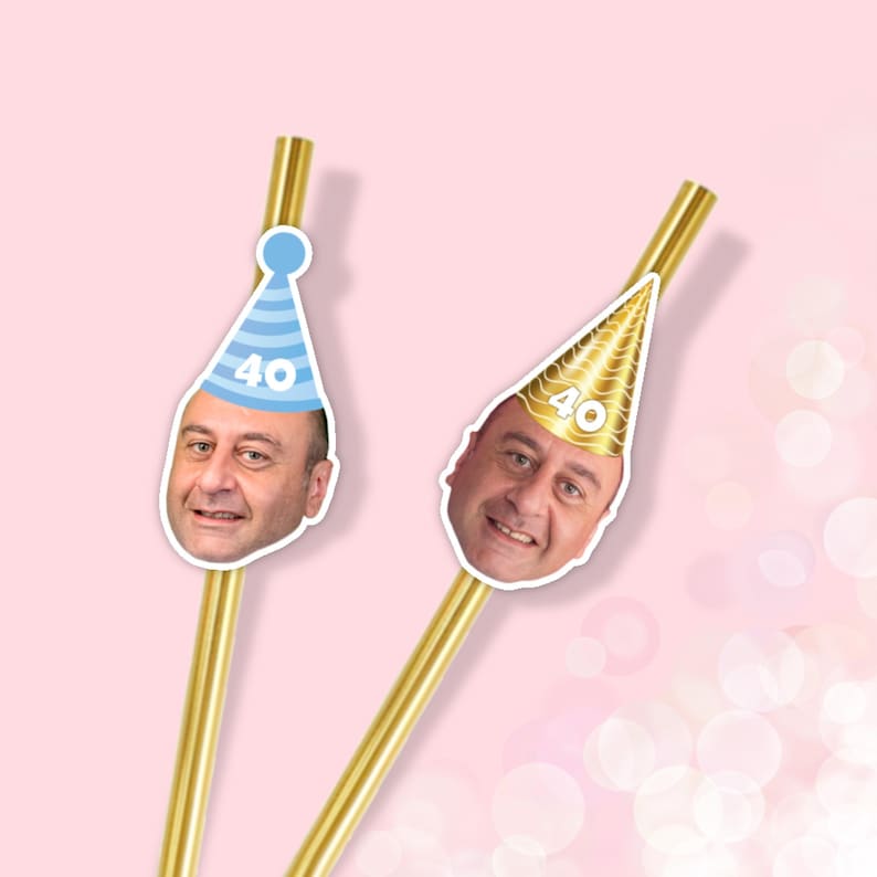 May include: Two gold-colored straws with attached face cutouts. One cutout features a man's face wearing a blue party hat with the number 40. The other cutout shows the same man wearing a gold party hat with the number 40. The background is a light pink color.