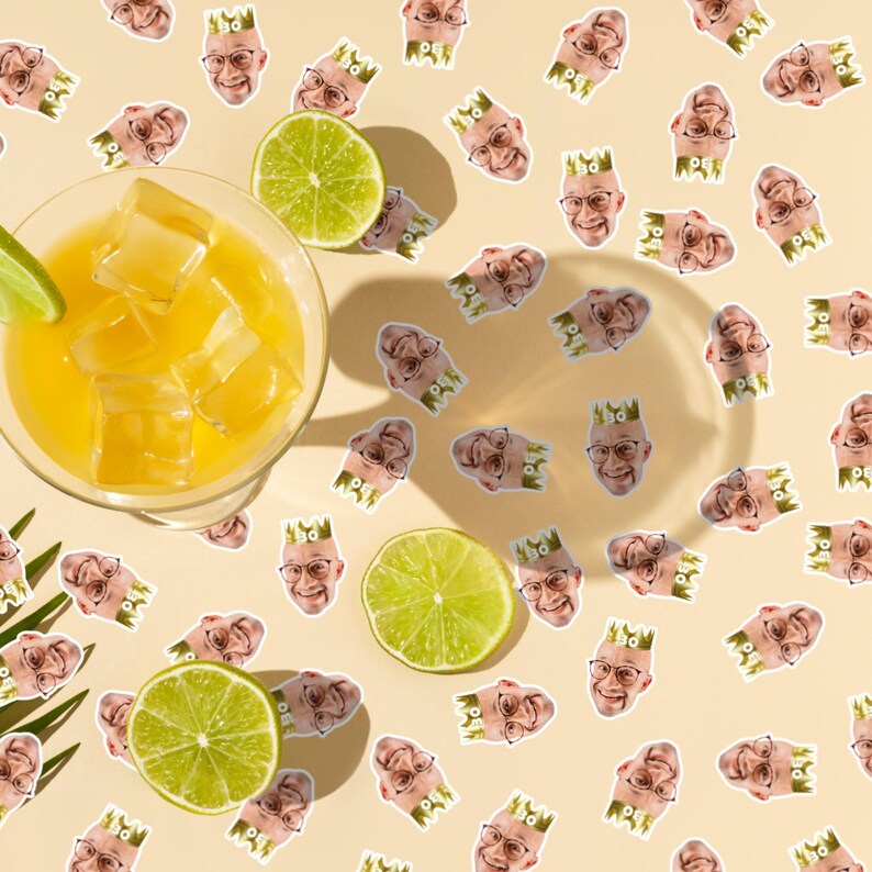 Puede incluir: Un c&oacute;ctel refrescante en un vaso transparente con cubitos de hielo y una rodaja de lima. La bebida est&aacute; rodeada de rodajas de lima y pegatinas de un hombre con gafas y una corona, todo sobre un fondo amarillo claro.