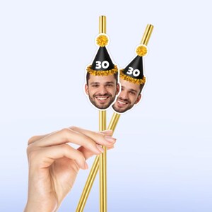 May include: Gold-colored straws with attached party hats featuring a smiling face and the number 30. The hats are black with a gold pom-pom and trim. The straws are held in a hand against a light blue background.