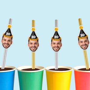 May include: Four colorful paper cups with straws featuring a smiling face wearing a party hat with the number 30. The straws are silver and gold, and the cups are red, yellow, green, and blue. The background is a light blue.