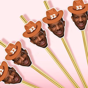 May include: Gold-colored straws with a smiling face wearing a brown cowboy hat with the number 32 printed on it. The straws are arranged diagonally against a pink background, creating a festive and celebratory theme.