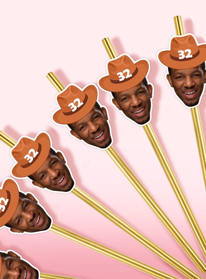 May include: Gold-colored straws with a smiling face wearing a brown cowboy hat with the number 32 printed on it. The straws are arranged diagonally against a pink background, creating a festive and celebratory theme.