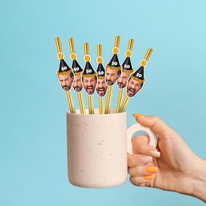 Puede incluir: Una taza rosa claro llena de pajitas doradas. Cada pajita tiene una tapa decorativa con la cara de un hombre que lleva un gorro de fiesta negro con el n&uacute;mero 50. El fondo es azul claro liso.