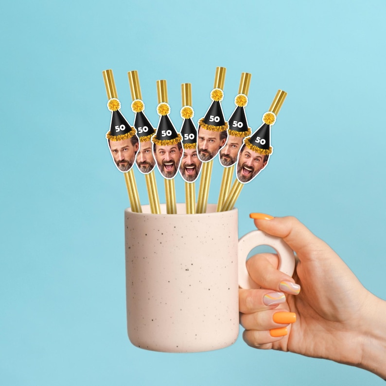 Puede incluir: Una taza rosa claro llena de pajitas doradas. Cada pajita tiene una tapa decorativa con la cara de un hombre que lleva un gorro de fiesta negro con el n&uacute;mero 50. El fondo es azul claro liso.