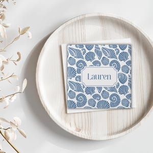 Könnte beinhalten: Eine quadratische Karte mit blauem Muschelmuster und dem Namen "Lauren" in einem weißen Rahmen, die auf einem hellen Holztablett liegt. Die Karte hat einen weißen Rand. Getrocknete Blumenäste befinden sich links.