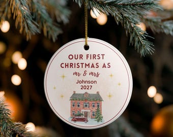 May include: A circular Christmas ornament with the inscription "OUR FIRST CHRISTMAS AS mr & mrs Johnson 2027". It displays a watercolour illustration of a house and car, and has a gold hanging string.