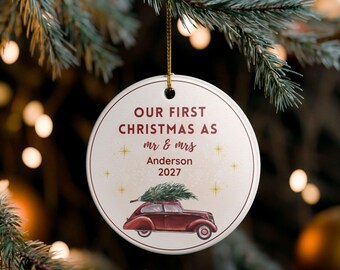May include: A circular Christmas ornament with the text "OUR FIRST CHRISTMAS AS mr & mrs Anderson 2027". A maroon car carrying a Christmas tree is depicted below the text. The ornament is suspended by a gold string.