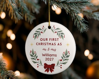 Puede incluir: Adorno navideño redondo blanco con la inscripción "OUR FIRST CHRISTMAS AS mr & mrs Williams 2027". El adorno presenta un lazo rojo, hojas verdes y bayas rojas. Una cuerda dorada está unida para colgarlo.