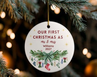 Puede incluir: Un adorno navideño de cerámica blanca con el texto "OUR FIRST CHRISTMAS AS mr & mrs Williams 2027". El adorno presenta un lazo verde, vegetación festiva y pequeñas medias.