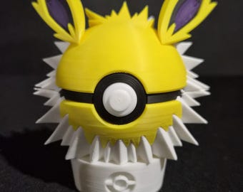 May include: A spherical yellow and white character figure with pointed ears, purple inner ear details, and a central black and white circular button design. The figure features a spiky white collar and rests on a white cylindrical base with a small circular emblem.