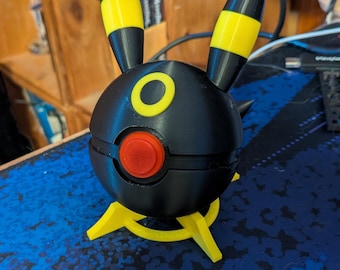 May include: A black and yellow Umbreon figure, a Pokémon character, with a round body, yellow accents, and pointed ears. The figure has a red button and yellow feet. The figure is on a blue surface.