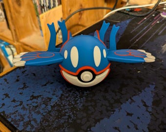 Może przedstawiać: Niebieska figurka Kyogre z białymi i czerwonymi akcentami. Figurka ma przypominające skrzydła wyrostki i spoczywa na niebiesko-czarnej wzorzystej powierzchni. To przedmiot kolekcjonerski.