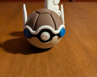 Może przedstawiać: Szczegółowa, trójwymiarowa figurka postaci z Pokemonów. Figurka jest głównie brązowa, biała i niebieska, z zaokrąglonym korpusem i stylizowanymi cechami. Ustawiona na ciepłej, drewnianej powierzchni.