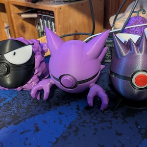 May include: Three 3D-printed Pokémon figures in black, purple, and dark purple. The black figure resembles a Poké Ball with white eyes. The purple figure is a Gengar. The dark purple figure has spikes and a red center.