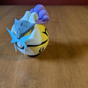 May include: A 3D-printed figurine featuring a stylized character with a white, yellow, and black body. It has blue and gray accents, including a star-shaped element and a headpiece. A purple cloud-like shape is on the back. The figurine is on a wooden surface.