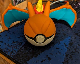 May include: An orange and white Poke Ball with Charizard wings and flame. The wings are teal and orange, and the flame is yellow and red. The figure is on a blue and black surface.