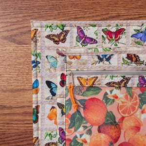 May include: A multi-compartment zippered pouch with a butterfly and orange fruit design. The pouch features a beige background with butterfly illustrations resembling postage stamps. The lower section displays oranges and flowers. An orange pull and a tree charm adorn the zipper.