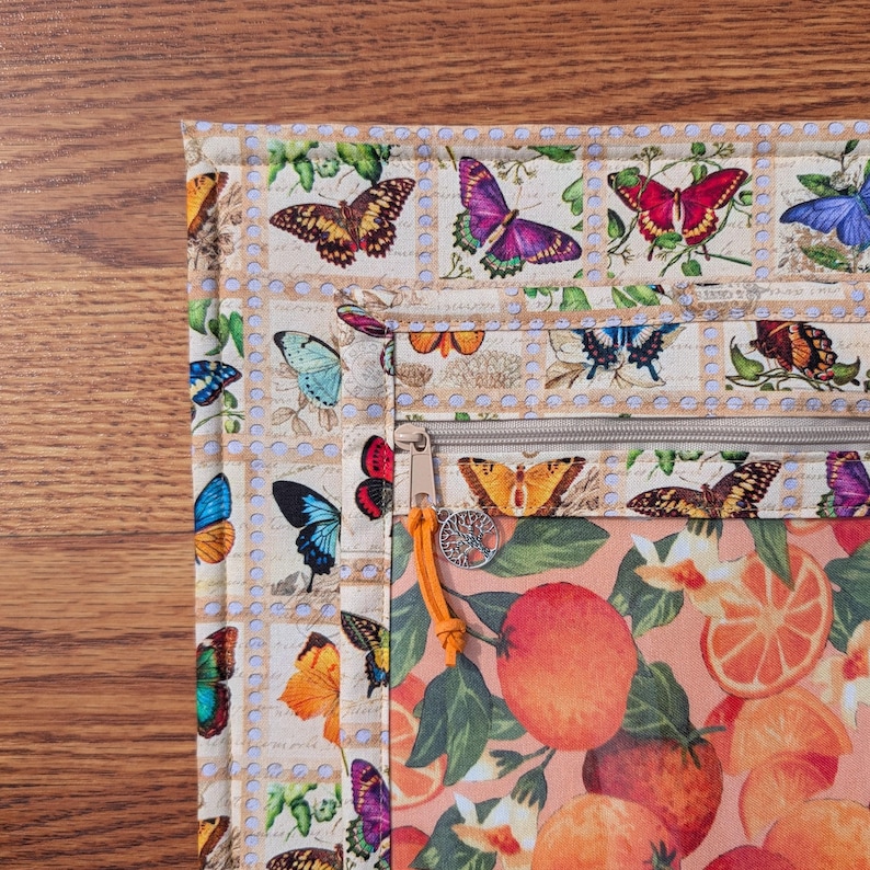 May include: A multi-compartment zippered pouch with a butterfly and orange fruit design. The pouch features a beige background with butterfly illustrations resembling postage stamps. The lower section displays oranges and flowers. An orange pull and a tree charm adorn the zipper.