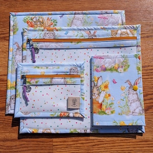 May include: A set of four fabric pouches with a light blue bunny print. The pouches vary in size and feature orange zippers, purple zipper pulls, and a small label. The bunny print includes rabbits, flowers, and Easter eggs.