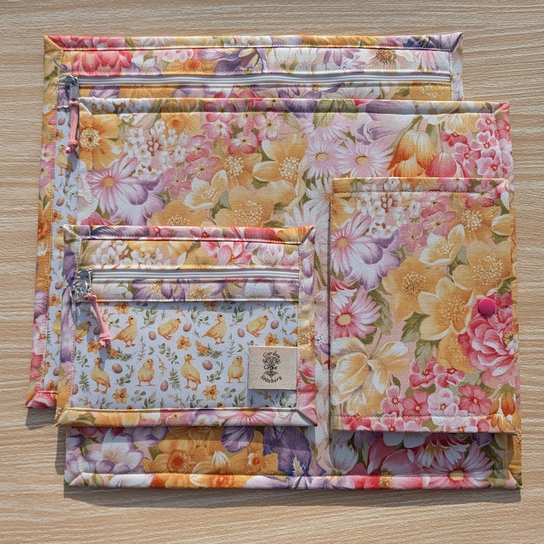Puede incluir: Un conjunto de bolsas de tela con estampado floral en varios tama&ntilde;os. Las bolsas presentan un patr&oacute;n de flores amarillas, rosas y moradas sobre un fondo claro. Una bolsa tiene un bolsillo transparente con un dise&ntilde;o de pollitos y huevos. Cremalleras y un cierre de bot&oacute;n rosa son visibles.