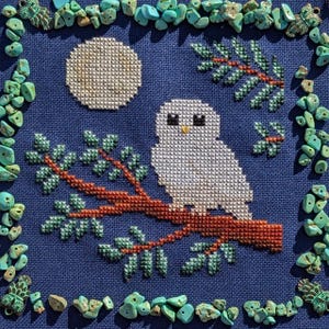 May include: Cross-stitch artwork of a white owl perched on a brown branch, set against a dark blue background with a full moon. Green leaves and turquoise stones frame the image, creating a nature-inspired design.