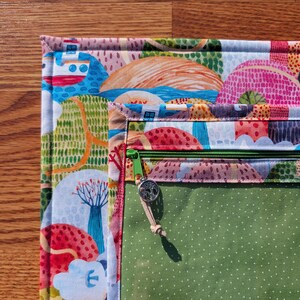 Puede incluir: Un estuche acolchado y colorido con un patr&oacute;n de paisaje caprichoso. Cuenta con una cremallera verde y un interior verde con lunares blancos. El dise&ntilde;o exterior incluye ilustraciones de casas, &aacute;rboles y barcos.