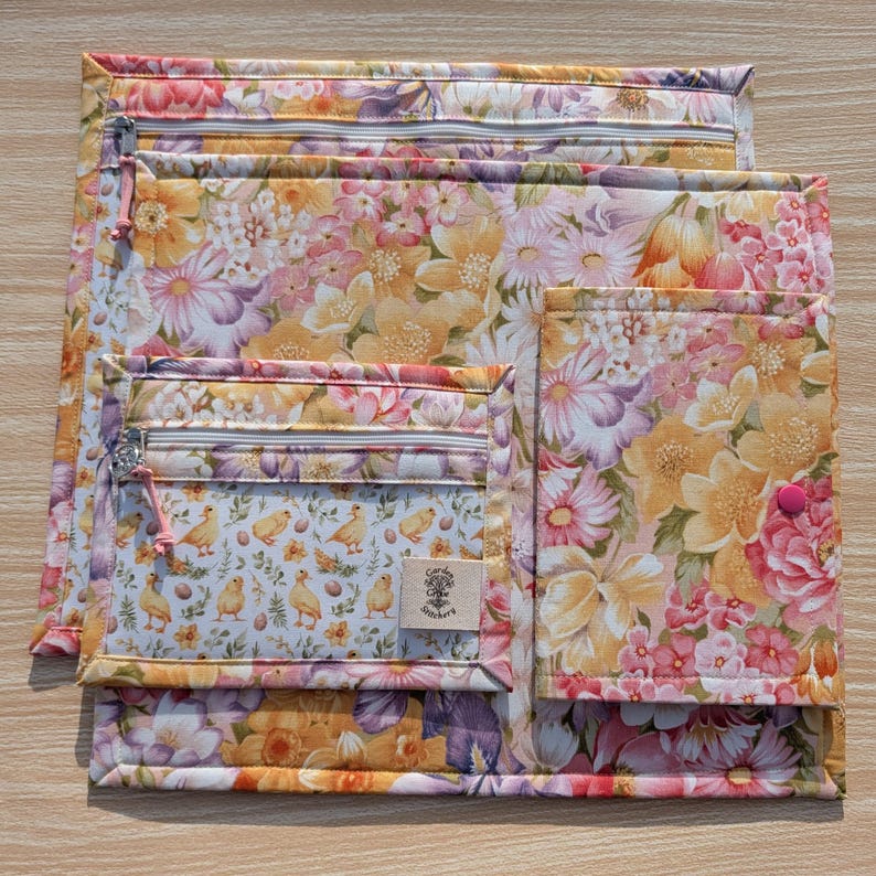 Puede incluir: Un conjunto de bolsas de tela acolchada con un estampado floral en tonos amarillos, rosas y morados. Las bolsas var&iacute;an en tama&ntilde;o y tienen cierres de cremallera. Una bolsa presenta un bolsillo m&aacute;s peque&ntilde;o con un patr&oacute;n de pollitos y huevos y una etiqueta peque&ntilde;a.