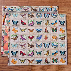 May include: Two beige quilted zipper pouches with a butterfly stamp design. The pouches feature a variety of colorful butterflies in shades of blue, red, orange, and purple. One pouch has a clear side with a fruit pattern.