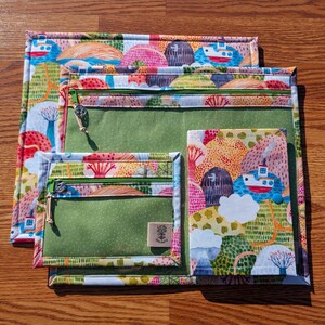 Puede incluir: Un conjunto de cuatro bolsas de tela con un colorido estampado de paisaje. Las bolsas tienen compartimentos verdes con cremallera y son de varios tama&ntilde;os, con un dise&ntilde;o de paisaje con barcos, &aacute;rboles y colinas en tonos rosa, azul y naranja.